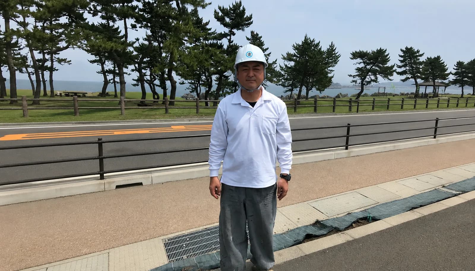 海沿いの道路に立つ、ヘルメットに白い作業着姿の代表 田中雅章