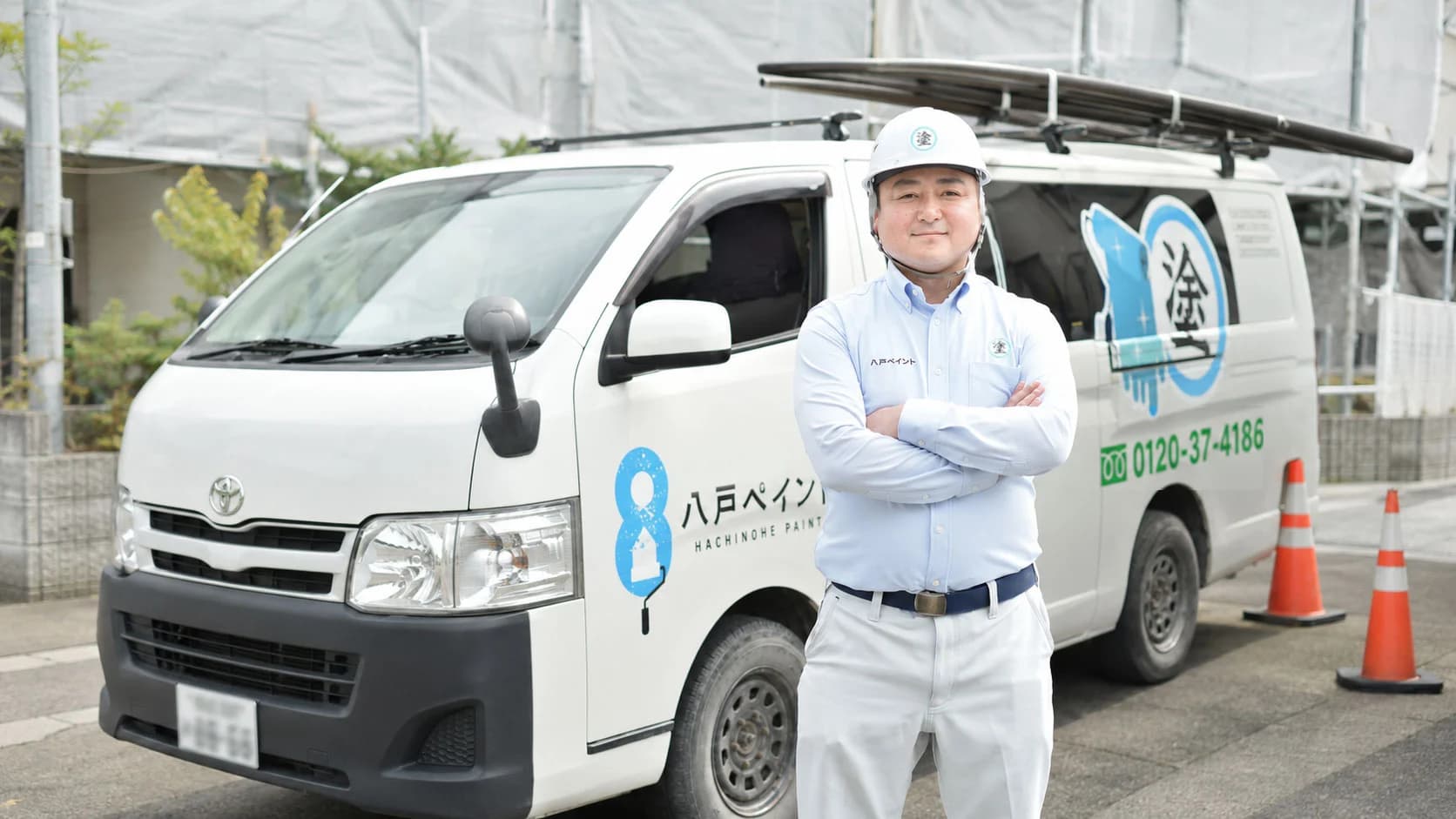 八戸ペイントのロゴを入れた社用車の前で腕を組む代表 田中雅章