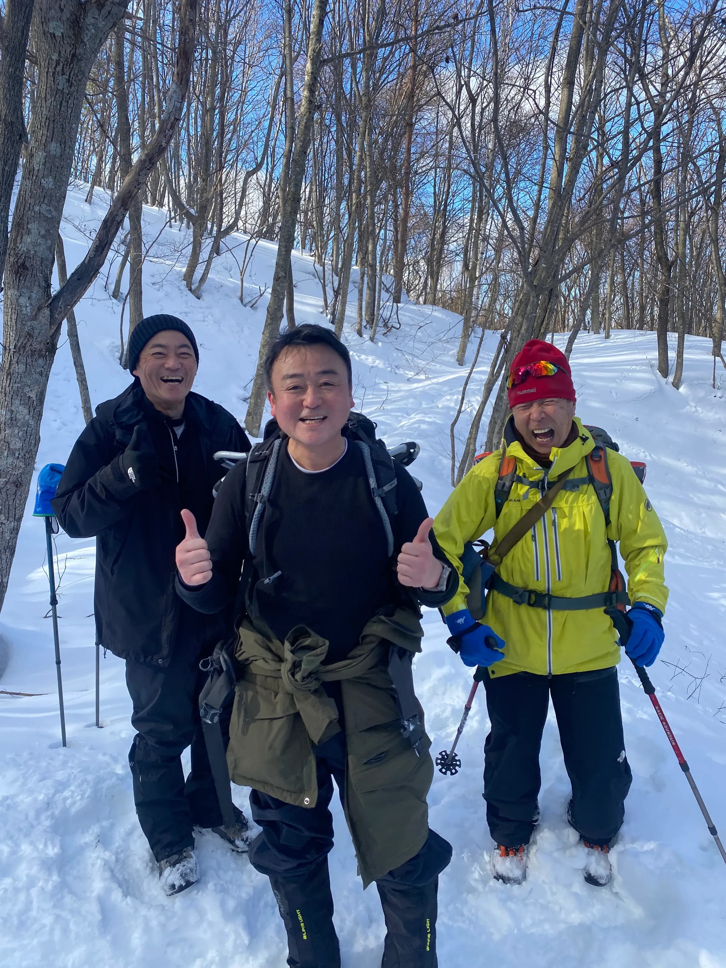 雪の林で、防寒着とスノーシューの装備で並び、親指を立てる3人の社員
