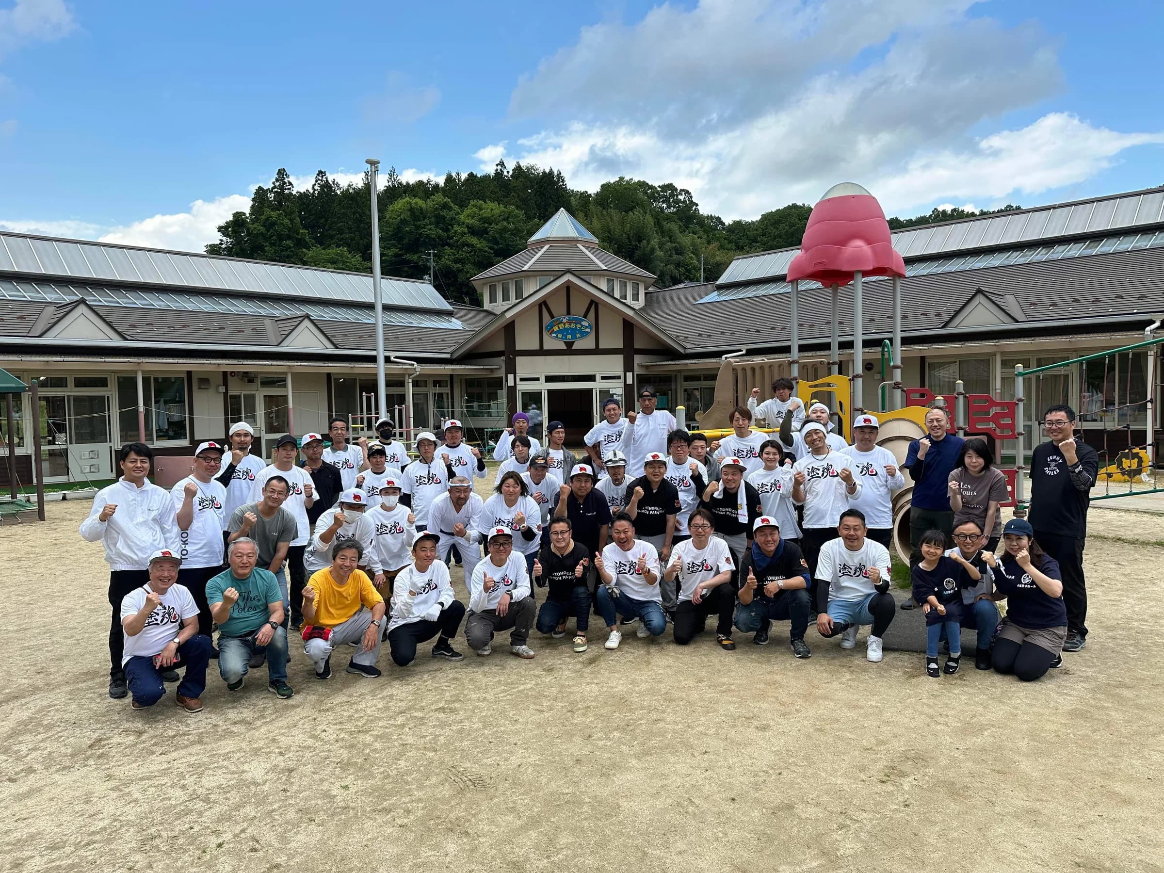 保育園の園庭で、塗魂ペインターズのTシャツを着た大勢の参加者が並んで写る集合写真