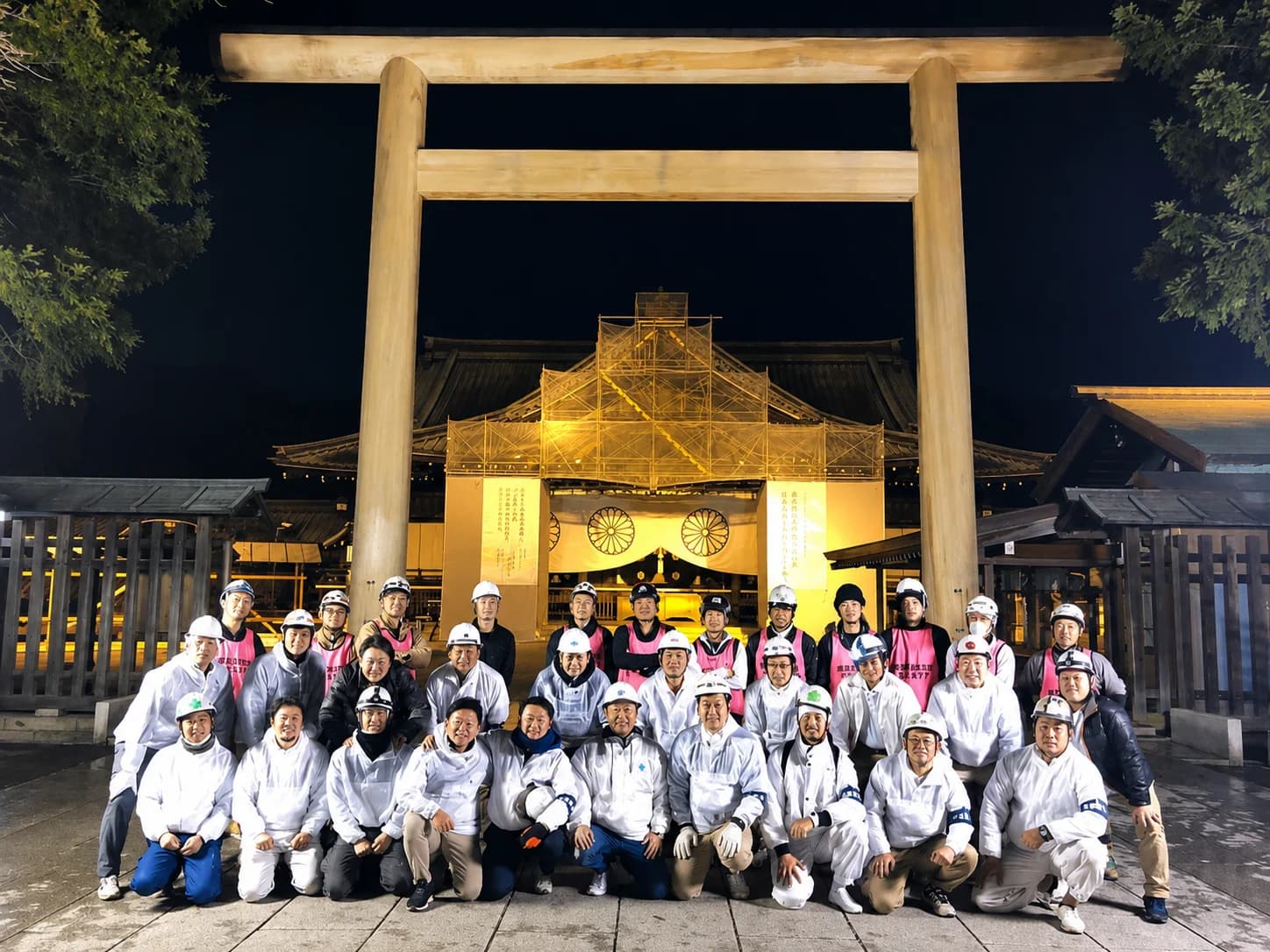 靖国神社の鳥居の前に、白い作業着とヘルメット姿で並ぶ参加者たちの夜の集合写真