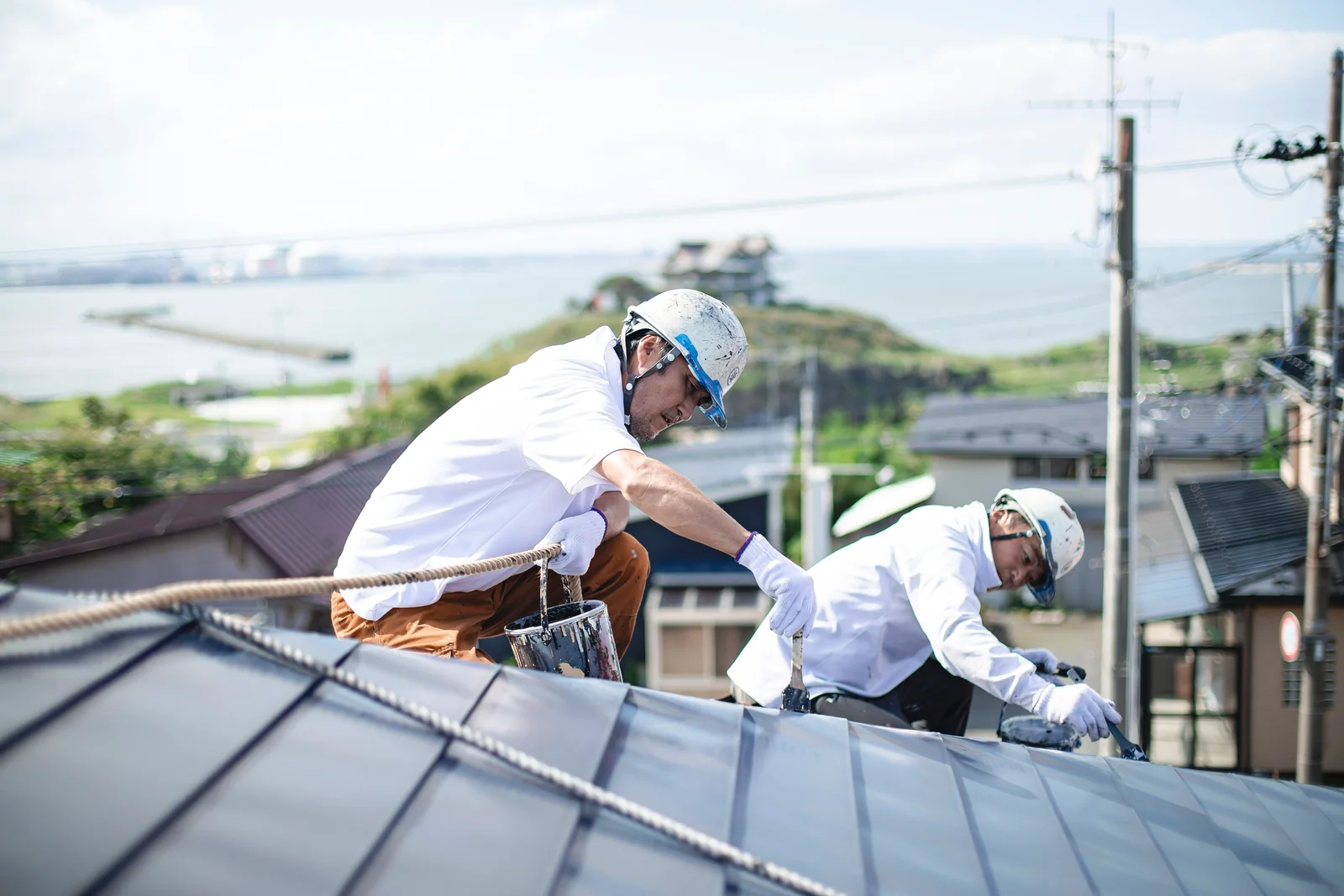 金属屋根の上で、ローラーを使って屋根を塗る2人の職人。奥に海と住宅街